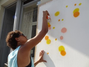 nancy is holding an aerosol can spraying yellow and orange dots on a white background