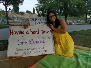Picture of Dee, in a yellow dress, smiling in a park on a green-yellow striped blanket with a sign "IG-Handle: pop.up.friend. Having a hard time? Come talk to me *smile*. Donations appreciated for helpful answers *heart*"