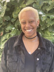 Potrait von Taye: Eine Schwarze Person lächelt mit blond gefärbten Haaren und Augenbrauen in einer schwarzen Jacke